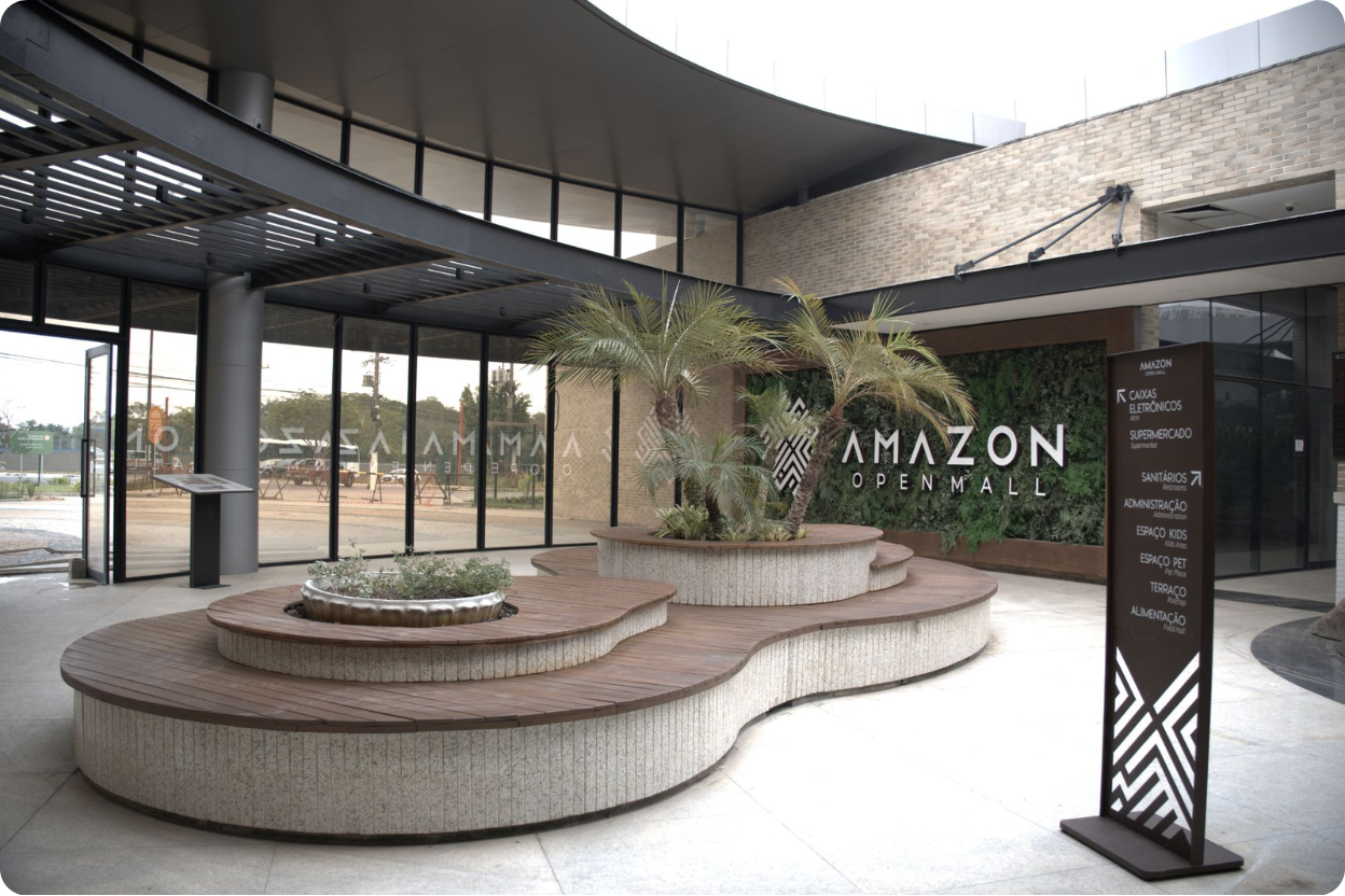 Para a última imagem deste conjunto (o Amazon Open Mall), aqui está uma sugestão de Alt Text focada em acessibilidade: "Fotografia da área interna e moderna do Amazon Open Mall em Manaus. O espaço possui design contemporâneo com teto alto de linhas curvas, paredes de tijolos claros e amplas janelas de vidro. No centro, há bancos de madeira em formatos orgânicos que circundam pequenos jardins com palmeiras. À direita, um totem informativo elenca serviços como supermercado e espaço pet, posicionado ao lado de uma parede com jardim vertical onde se lê o logotipo 'AMAZON OPEN MALL'.